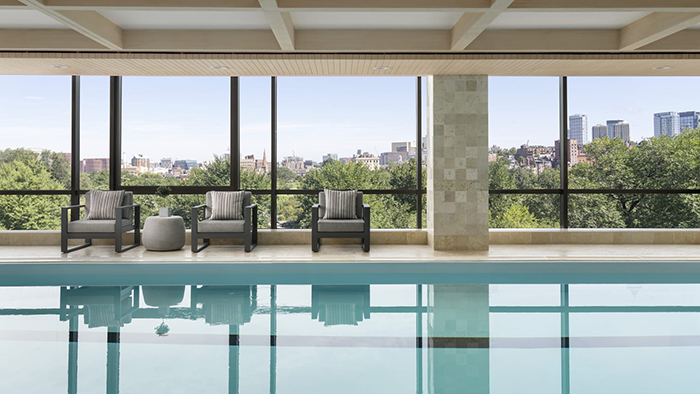 An indoor pool at the Four Seasons Hotel Boston with a large window facing a park with the city skyline just beyond the treetops.