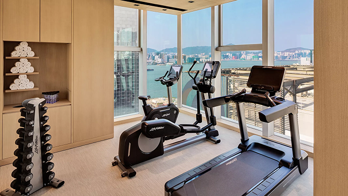 The fitness center at the AKI Hong Kong - MGallery hotel. A treadmill, an elliptical, and a stationary bike face a window with a view of the city.