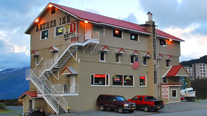 The exterior of the Anchor Inn Hotel in Whittier, Alaska. Two cars, a couple bikes, and a boat are parked outside.