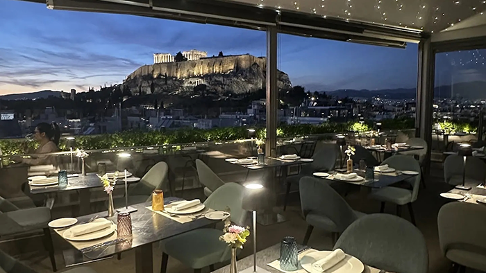 A restaurant on top of the Athens Gate Hotel. The room is dimly lit, and through the window is an incredible view of the Acropolis.
