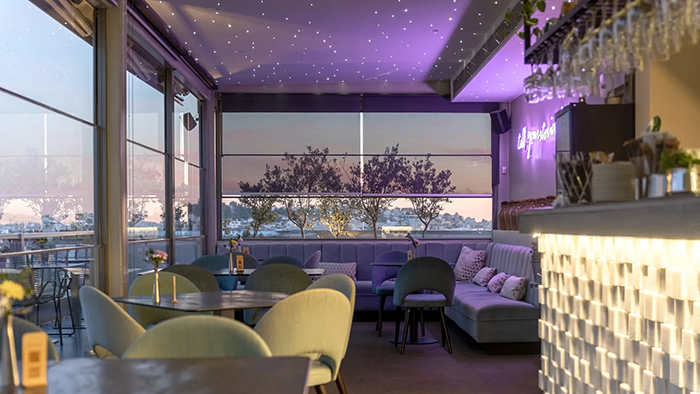 A bar on top of at the Athens Gate Hotel, photographed at dusk. Purple lighting from an LED sign creates a romantic atmosphere.