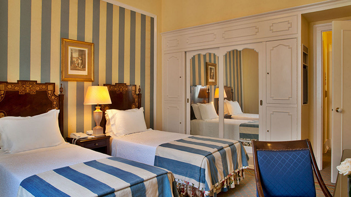 A guest room at the Avenida Palace hotel in Lisbon, Portugal. There are two twin beds with striped comforters that match the wallpaper.