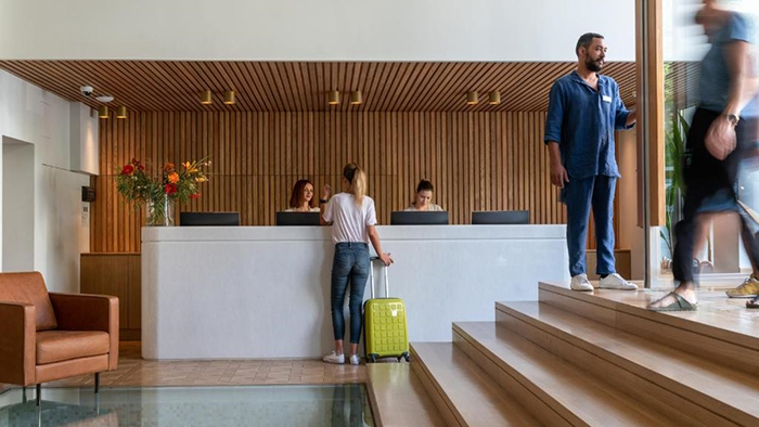 Guests passing through the lobby of the Coco-Mat Athens BC hotel. Two staff members stand behind the front desk while one opens the front door allowing two people to enter the building.