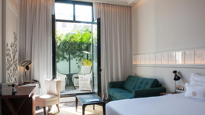 A guest room inside the Autograph Collection Cotton House Hotel. Two glass doors are open and lead to a terrace with seating and plant life.