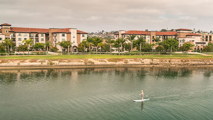 A view of the Homewood Suites by Hilton San Diego Airport-Liberty Station hotel from the water, where a man can be seen paddleboarding.