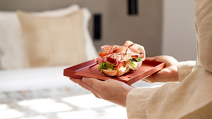 Room service being delivered to a guest room at in[n]Athens hotel. A staff member in a beige shirt carries an open faced sandwich with prosciutto, lettuce, tomato, and a balsamic vinaigrette drizzle.