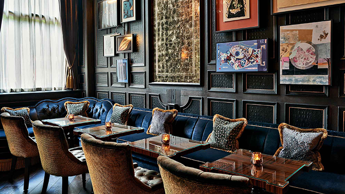 A comfortable-looking seating area inside The Kimpton Fitzroy London Hotel. The wall behind the blue velvet bench features numerous pieces of artwork.