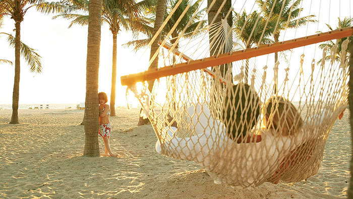 A couple relaxes in a beach hammock at the Lago Mar Beach Resort & Club while their child leans against a palm tree.