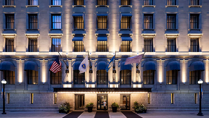 An exterior shot of The Langham Hotel in Boston. A staff member prepares to open a door underneath an awning with 