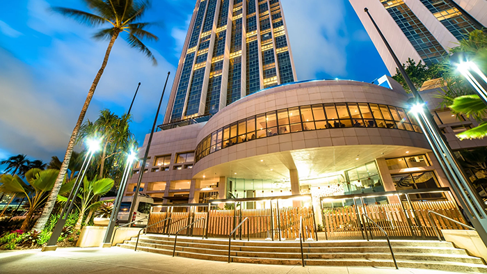 The exterior of the Prince Waikiki, a Honolulu Luxury Hotel. An extremely tall palm tree is visible to the left of the building.