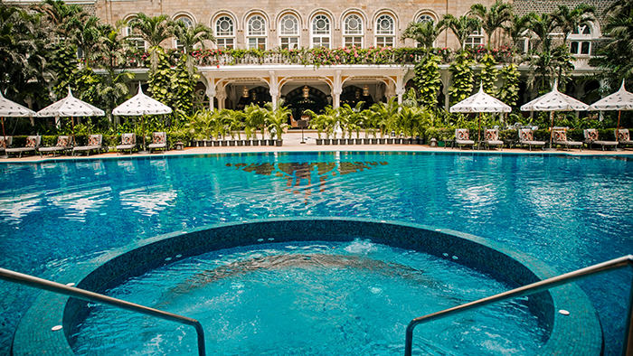 The Taj Mahal Palace Hotel pool. Two rows of lounge chairs sit under umbrellas at the edge of the water.