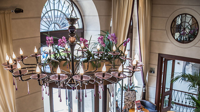 A hanging light fixture featuring several purple flowers inside the Victoria Hotel Letterario. A man can be seen sitting on a couch at the bottom of the image.
